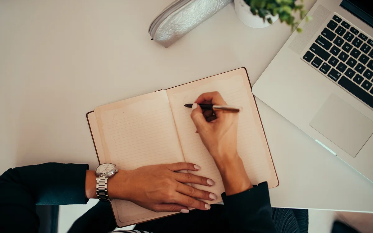 A person writing in a notebook beside a laptop at a modern workspace.
